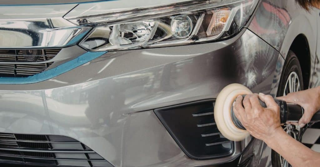 A professional technician carefully buffs the grey front bumper of a Ford vehicle near the fog light housing using a handheld orbital polisher.