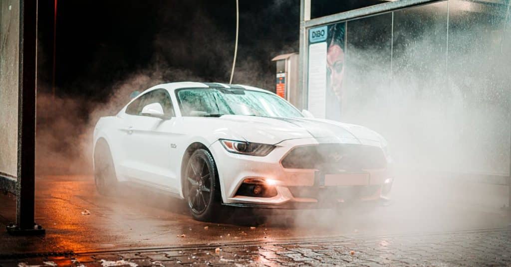 A white Ford Mustang GT with black racing stripes sits in an illuminated wash bay surrounded by steam and water mist after a professional cleaning.