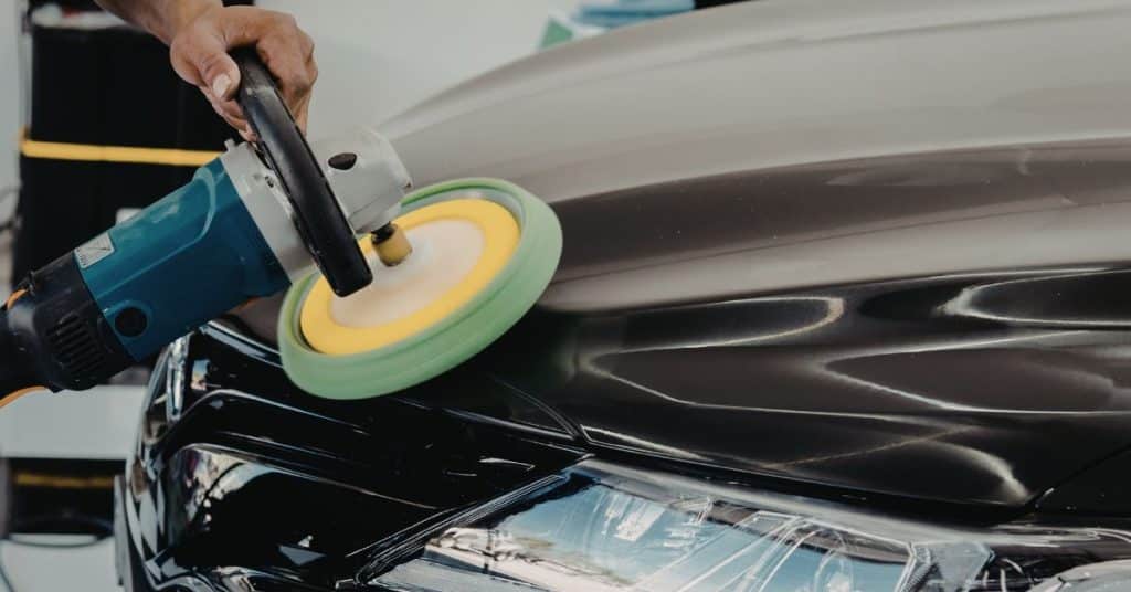 A close-up view of a technician using a high-speed rotary polisher with a yellow and green pad on the black hood of a vehicle to restore its showroom shine.